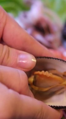 El, buğulanmış deniz tarağı kabuğundan eti açar ve çıkarır. İçindeki taze eti ortaya çıkarır. Arka planda toplanan bir kase midye eti de deniz ürünlerinin hazırlanma anını betimler..