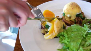 Benedict yumurtalı, salatalı, meyveli ve portakallı sağlıklı kahvaltı yapan kişi açık kafede. Taze, besleyici sabah yemeği konsepti. Ağır çekim. Yüksek kalite 4k görüntü