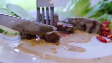 Beyaz, kavrulmuş bir yüzey kenarındaki ızgara bifteği kesen bir çatal ve bıçağın yakın plan çekimi ve sulu doku bifteğin zengin lezzetini ve hassasiyetini ön plana çıkarıyor. Yüksek kalite 4k görüntü