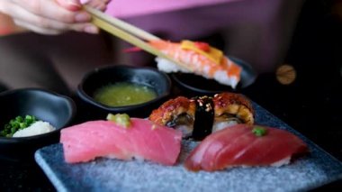 Bir el ustalıkla Nigiri suşisini yemek çubuklarıyla, dramatik stüdyo ışıklarının altında, siyah bir arkaplan eşliğinde, düzgün düzenlenmiş bir tabaktan seçiyor. Yüksek kalite 4k görüntü