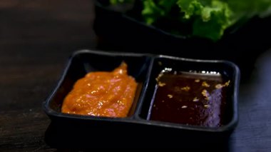 Sarımsak ve kırmızı biber parçacıkları içeren koyu bir sosla dolu küçük metal bir kabın yakın çekim görüntüsü. Konteynır tereyağı ve taze marulun yanına bir tepsiye konur. 