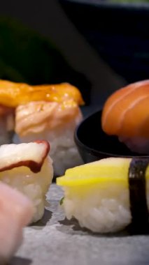 Nigiri, sashimi ve tamago 'yu da içeren beyaz bir tabak suşi, parlak ve hatta aydınlık arka planda siyah bir zemine karşı sorunsuzca döner. Yüksek kalite 4k görüntü