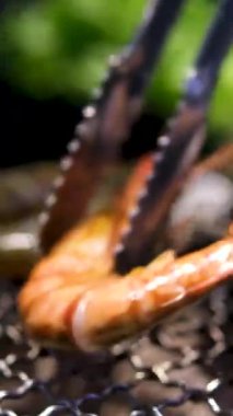 Izgarada kızaran lezzetli karidesler. Metal bir ızgarada taze karides ızgara ediliyor. Maşayla el, deniz mahsullerini sıcak kömürlerin üzerinde döndürür, yaz barbeküsünün canlı renklerini ve dokularını yakalar.