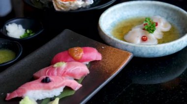 Suşi, somon, ton balığı, deniz tarağı ve somon yumurtası gibi çeşitli suşiler, garnitürlerle dolu bir tabakta sunuluyor, stüdyo ışıkları altında düzgün bir şekilde dönüyor. Yüksek kalite 4k görüntü