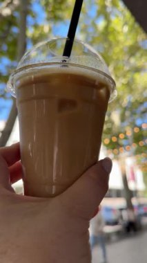 Yaz günü şehir ortamında bir kadının elinde buzlu kahve bardağını samanla tutarken yakın plan. Bulanık şehir arkaplanının aksine, odak noktasında soğuk, ferahlatıcı bir içecek. Çekim.