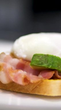 Avokado dilimli pastırmalı ve yumurtalı sandviç siyah eldivenli şefi beyaz tabakta bir dilim ekmeğin üzerine yerleştirir.