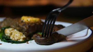 Beyaz, kavrulmuş bir yüzey kenarındaki ızgara bifteği kesen bir çatal ve bıçağın yakın plan çekimi ve sulu doku bifteğin zengin lezzetini ve hassasiyetini ön plana çıkarıyor. Yüksek kalite 4k görüntü