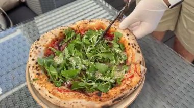 Strachatella peynirli jambonlu pizza, domates ve roka yapraklı. Tahta masada dönüyor. Pizzayı makasla kes.