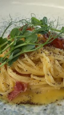 Otantik İtalyan carbonara makarnası kremasız, altın çıtır pastırma ve beyaz tabakta taze mikroyeşiller kameranın önünde yavaşça dönüyor..