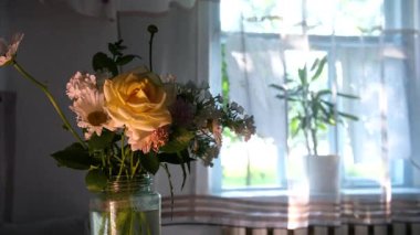 Papatya buketi ve güller kır evindeki ahşap pencerenin yanında durur, rüzgar perdeleri yavaşça hareket ettirir, sıcak güneş ışığı kırsal bölgede huzurlu bir yaz atmosferi yaratır..