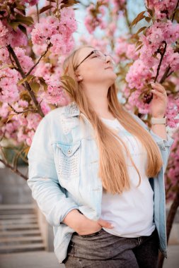 Görüntü, baharda açık havada çiçek açan pembe bir sakura ağacının yanında poz veren sarışın bir kızın portresini gösteriyor..