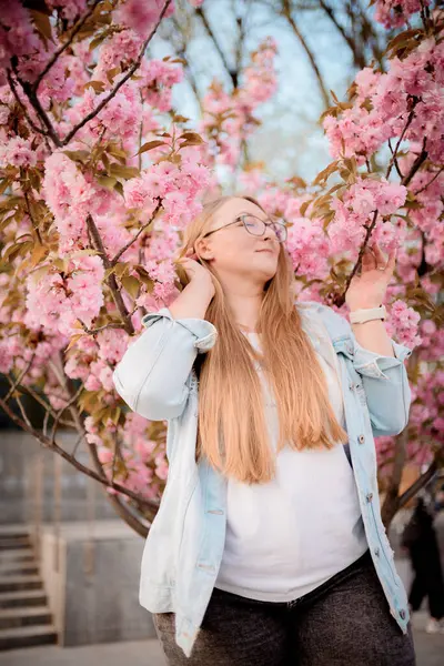 Görüntü, baharda açık havada çiçek açan pembe bir sakura ağacının yanında poz veren sarışın bir kızın portresini gösteriyor..