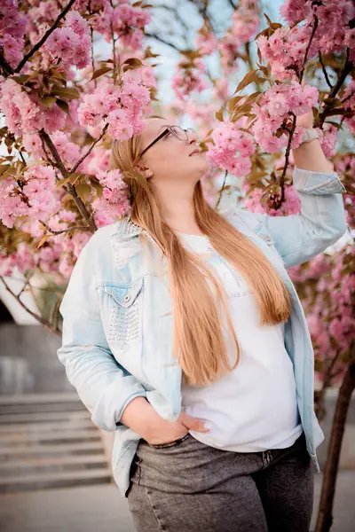 Görüntü, baharda açık havada çiçek açan pembe bir sakura ağacının yanında poz veren sarışın bir kızın portresini gösteriyor..