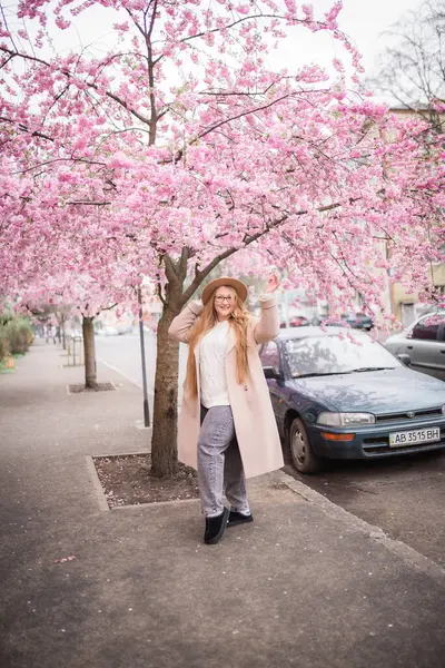 Fotoğrafta sokakta duran bir kız görebilirsiniz. Pembe bir sakura ağacı ve yeşil bir araba..