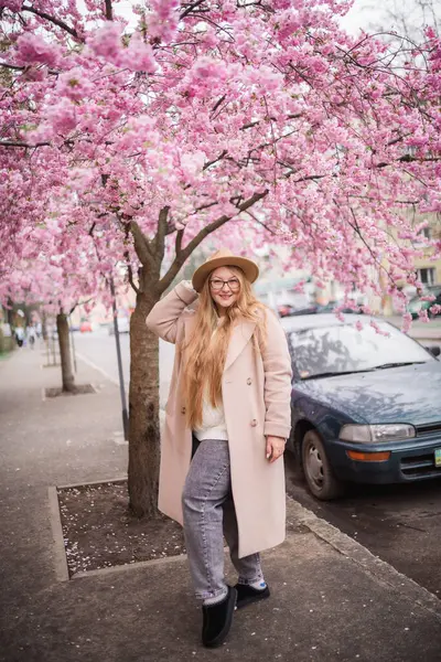 Fotoğrafta sokakta duran bir kız görebilirsiniz. Pembe bir sakura ağacı ve yeşil bir araba..