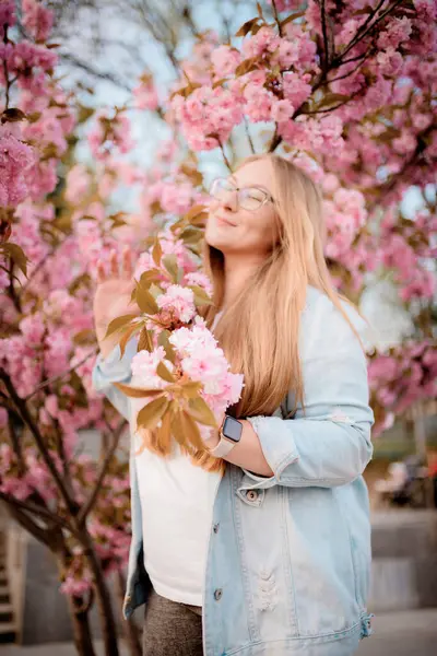 Görüntü, baharda açık havada çiçek açan pembe bir sakura ağacının yanında poz veren sarışın bir kızın portresini gösteriyor..
