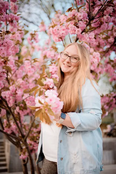 Görüntü, baharda açık havada çiçek açan pembe bir sakura ağacının yanında poz veren sarışın bir kızın portresini gösteriyor..