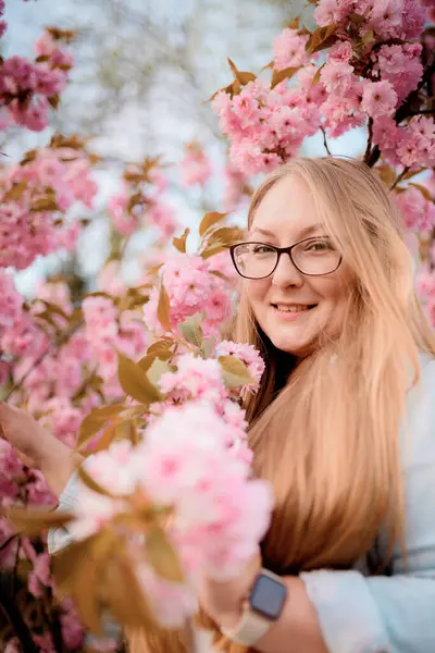 Görüntü, baharda açık havada çiçek açan pembe bir sakura ağacının yanında poz veren sarışın bir kızın portresini gösteriyor..