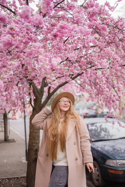 Fotoğrafta sokakta duran bir kız görebilirsiniz. Pembe bir sakura ağacı ve yeşil bir araba..