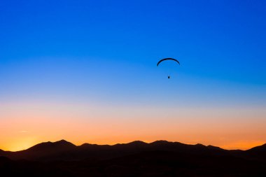 Bulutsuz mavi ve turuncu gökyüzüne ve dağların siluetine karşı bir paraglider silueti.