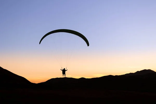 Bulutsuz mavi ve turuncu gökyüzüne ve dağların siluetine karşı bir paraglider silueti.