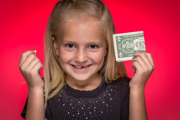 A girl with missing tooth holding removed tooth and dollar bill against a red background
