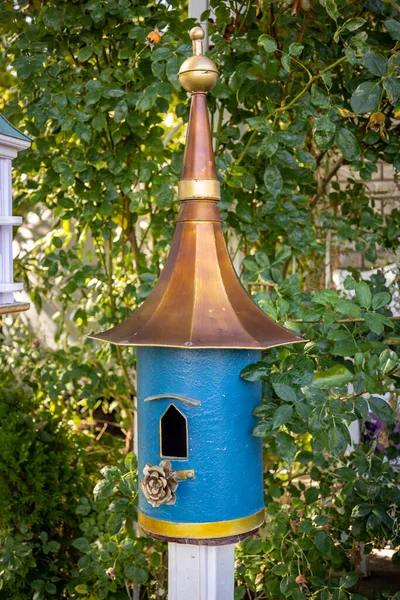 A bright blue birdhouse with a pointed copper roof mounted on a white post in a leafy garden in the day