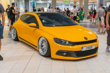 Canlı sarı bir Volkswagen GTI spor arabası kapalı bir otomobil fuarında sergileniyor, özel tekerlekleri ve pazar sonrası modifikasyonları sergileniyor.