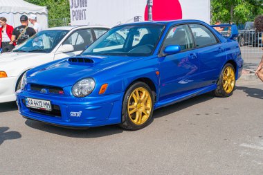 Mavi bir Honda Civic Tipi R, altın renkli jantlar ve beyaz bir plaka ile asfalt bir zemine park edilmiş, diğer arabalar ve bir çadırla çevrili. C Kyiv, Ukrayna 06-2024