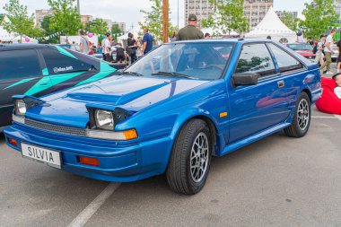 Modifiye edilmiş mavi bir araba halka açık bir alanda yüzeyinde parlak yansımaları olan hafif bir açıyla park edilmiş durumda. Araç Ukrayna 06-06-2024 'e ait.