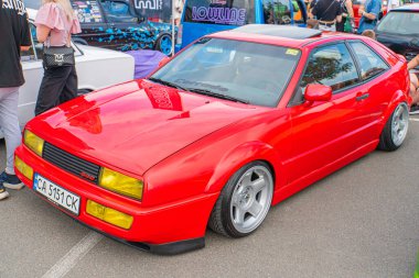 A red sports car with a modified performance appearance is parked on an asphalt surface, surrounded by other vehicles and people in the background, su Kyiv, Ukraine 06-06-2024 clipart