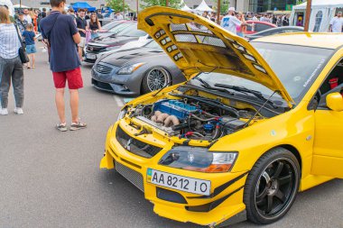 Kaputu açık, modifiye edilmiş sarı bir araba motorunu ortaya çıkarıyor. Piyasa tekerlekleri, yan etekler, çıkartmalar ve çıkartmalarla süslenmiş. İnsanlar Kyiv 'i, Ukrayna 06-06-2024' ü suretliyor.