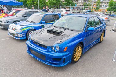 Mavi bir Subaru Impreza, kalabalık bir açık hava otomobil fuarı etkinliğinin merkezinde, diğer özelleştirilmiş araçlarla çevrili, Yiv, Ukrayna 06-06-2024 ile büyük bir şehir otoparkında yer alıyor.