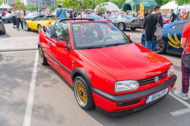 Canlı kırmızı bir hatchback arabası park halinde duruyor, yan camları kısmen açık, sarı ve siyah jantlar kırmızı gövdeye zıt. Bulanık Kyiv, Ukrayna 06-06-2024