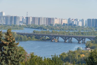 Dinyeper üzerindeki Kiev köprüsü. Nehrin üzerindeki köprüde giden arabalar. Büyük şehrin ve plajın arka planına karşı. Kiev, Ukrayna 10-29-2024.