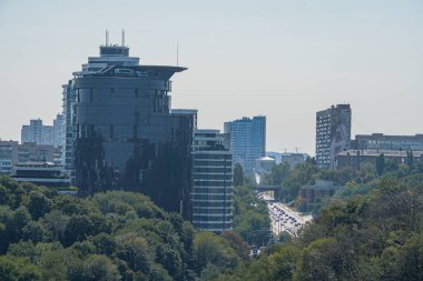 Görüntü, ön plandaki yemyeşil ağaçlar tarafından çerçevelenmiş kentsel bir manzara gösteriyor. Camdan ve metalden yapılmış binalar Kyiv, Ukrayna 06-06-2024