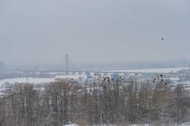 Ön planda karla kaplı dalları olan çıplak ağaçları gösteren bir kış manzarası, muhtemelen orta zemindeki küçük bir topluluk veya sanayi bölgesi Kyiv, Ukrayna 01-14-2024