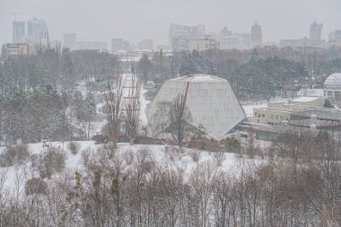 Wintry cityscape with snow-covered landscape Buildings, including a dome structure, visible in the distance Leafless trees and walkwayroad in foreground Overcast sky, Kyiv, Ukraine 01-14-2024 clipart