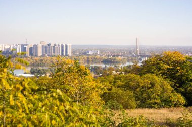 Sonbahar manzarasının panoramik görüntüsü, yoğun sarı-turuncu ağaç örtüsü, yansıtıcı su kütlesi kentsel alanı ve açık mavi gökyüzü altındaki doğal manzarayı ayırıyor.