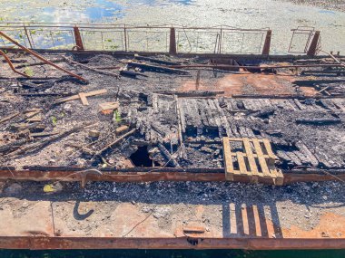 Şiddetli hasarlı bir tekne enkazı, yanmış ve yanmış, nilüferlerle dolu bir su kütlesinin etrafına saçılmış enkaz parçaları.