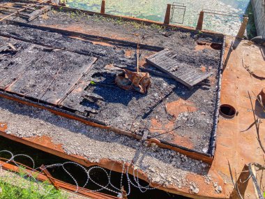 Image depicts a destroyed building amidst debris, with growing vegetation and a wire fence nearby The background features a calm body of water The image suggests a post-disaster scene without text clipart