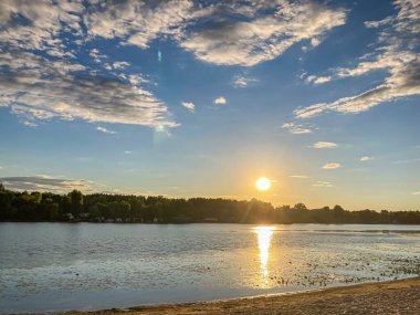 Deniz kenarında sakin bir gün batımı sahnesi, hafif dalgalı sakin sular, kıyı şeridinde bitki örtüsü, parıldayan güneş ışığı yansıması, huzurlu ve sakin bir atmosfer.