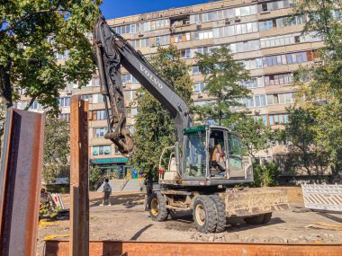 Yeşil ve siyah kazıcı, enkaz yığınları, devam eden yıkım veya yenileme çalışmaları, binalarla endüstri geçmişi ve uzak bir ağaç olan Kyiv, Ukrayna, 06-15-2024