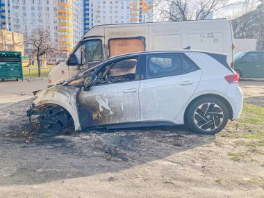 Kentsel bir ortamda yanmış, hasarlı beyaz bir araba, tanımlanamayan bir bina ve kısmen Ukrayna, Kyiv 'de görülebilen başka bir araç ile, herhangi bir insan varlığı veya müdahalesi belirtisi yok, 03-15-2024