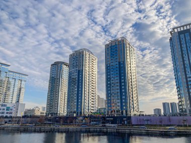 Sakin bir şehir manzarasında modern, mavi-beyaz binalar bulutlu bir gökyüzü altında sakin bir şehir limanına yansıyor. Fotoğraf mimariye ve Kyiv, Ukrayna, 03-15-2024 'e odaklanıyor.