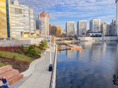 Bir yürüyüş yolu, banklar ve yemyeşil yemyeşil bir nehir manzarası binalarla çevrili bulutlu gökyüzünü yansıtıyor S Kyiv, Ukrayna, 03-15-2024