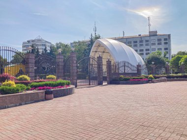 Gündüz açık hava manzarası, eşmerkezli tuğla daireli asfalt alan, yeşil ve kırmızı santralli alçak duvar, parka veya umumi alana açılan giriş kapısı, karmaşık metal işi Kyiv, Ukrayna, 06-15-2024