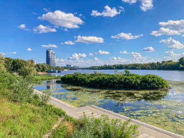 Beton bir yürüyüş yolu, nilüferler ve köprü altlığı ile huzurlu bir göl manzarası. Doğa ve insan yapımı yapılar bu huzurlu ortamda uyum içinde yaşıyorlar. Kyiv, Ukrayna, 06-15-2024