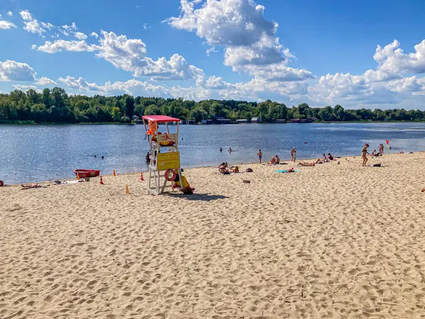 Kırmızı-sarı cankurtaran standının olduğu canlı bir sahil sahnesi, dinlenen kumsalcılar, mavi-yeşil sular, kıyıdaki botlar, açık gökyüzü, ve sabahın erken saatlerinde beliren doğal ışık, ya da I Kyiv, Ukrayna, 06-15-2024