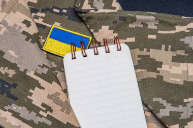 Askeri not defteri. Ukrayna bayrağı. Kamuflaj arka planında askeri temalı kişisel eşyalar serili. a